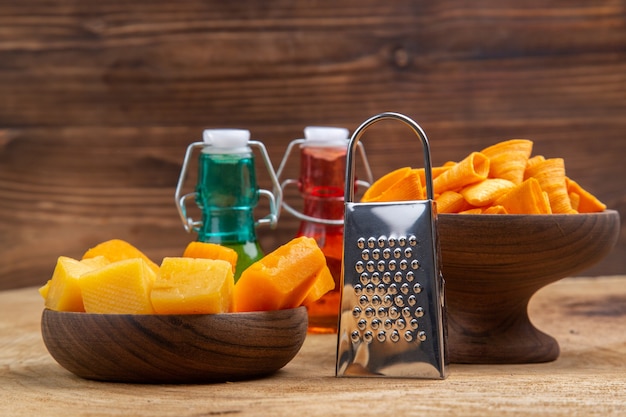 Front view slices of cheese and chips in bowls grater red and green bottles on brown isolated surface