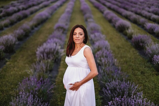 Front view of smiled brunette caucasian pregnant woman dressed in white dress, touching belly in lavender field.