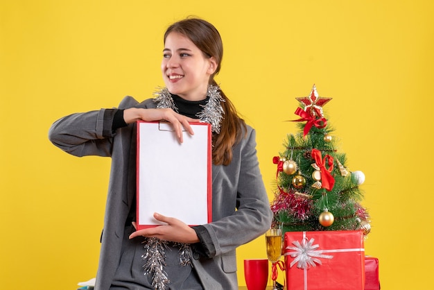 Free Photo front view smiled girl holding documents standing near xmas tree and gifts cocktail