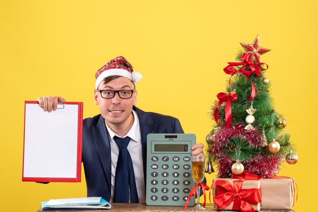 Front view of smiled man with santa hat sitting at the table near xmas tree and presents on yellow wall