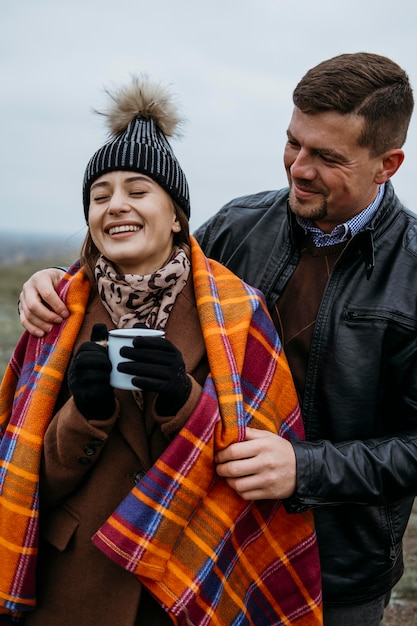 Front view of smiley couple outdoors with blanket and warm drink