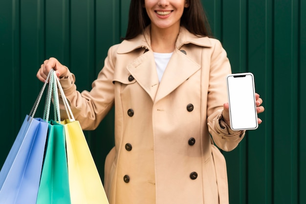 Front view of smiley woman holding up smartphone and shopping bags