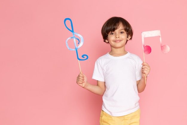 A front view smiling adorable boy with colorful signs in white t-shirt on the pink desk