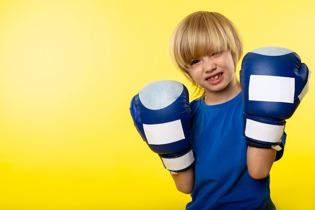 Free photo a front view smiling blonde boy posing boxing in blue boxing gloves on the yellow wall