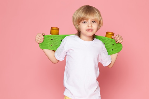 Free photo a front view smiling blonde boy in white t-shirt holding green skateboard on the pink space