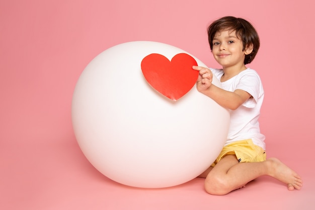 A front view smiling cute boy in white t-shirt playing with white ball on the pink desk