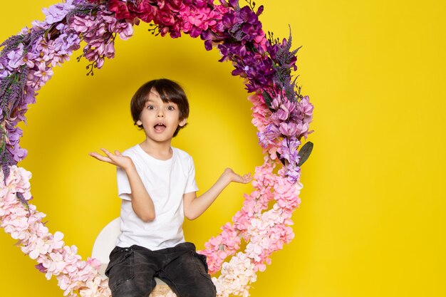 A front view smiling cute boy in white t-shirt sitting on the flower made stand on the yellow desk