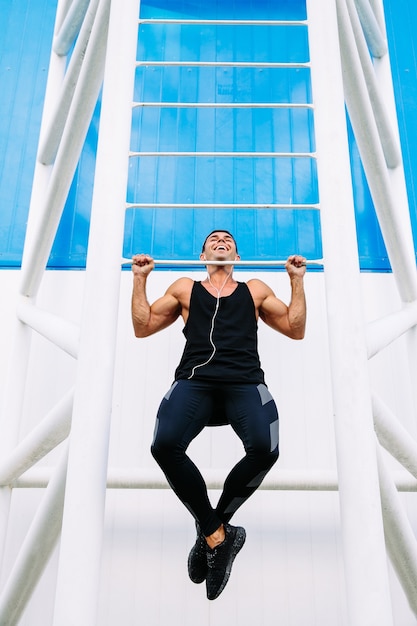 Front view of smiling handsome muscular guy doing pull ups, wearing black sportswear.