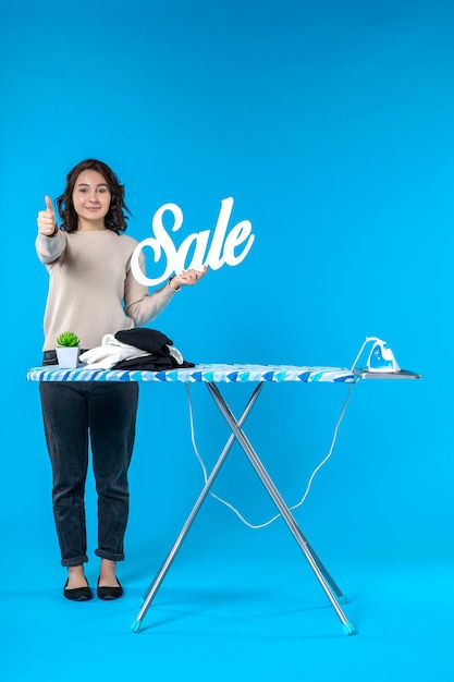 Front view of smiling happy young woman standing behind the ironing board and showing sale icon making ok gesture on blue wall