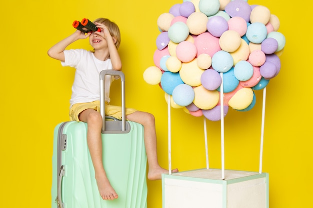 Free photo front view smiling kid blonde haired sitting on the blue bag along with colorful air balloons on yellow floor