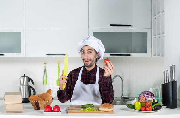 Front view of smiling male chef holding tomato and knife in the kitchen