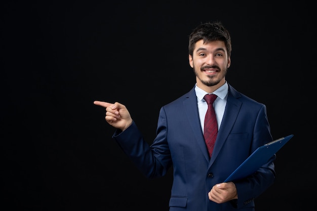 Front view of smiling male office worker in suit holding documents and pointing something on the right side on isolated dark wall