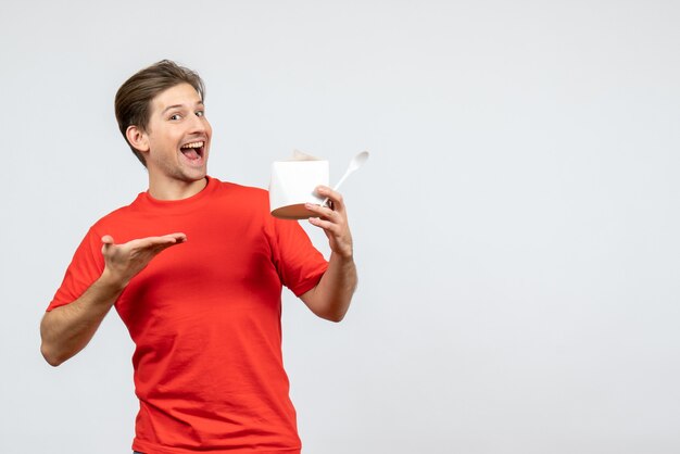 Front view of smiling young guy in red blouse pointing paper box on white background