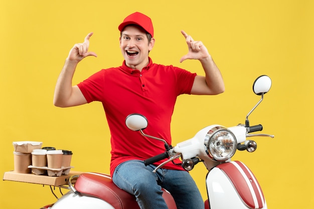 Front view of smiling young guy wearing red blouse and hat delivering orders making exact something on yellow background