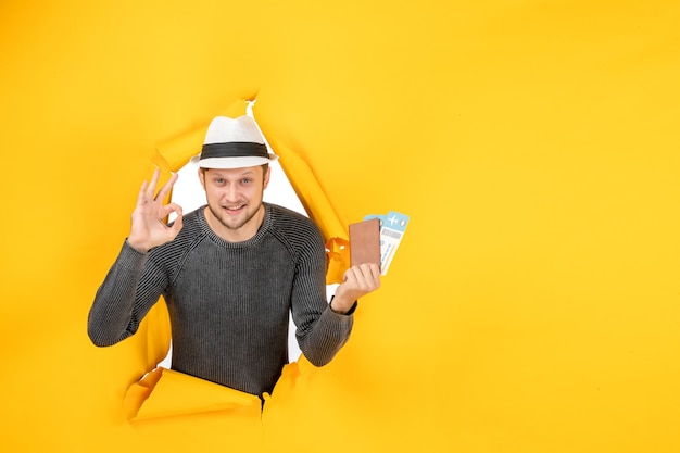 Free Photo front view of smiling young man with a hat holding foreign passport with ticket and making eyeglasses gesture in a torn on yellow wall