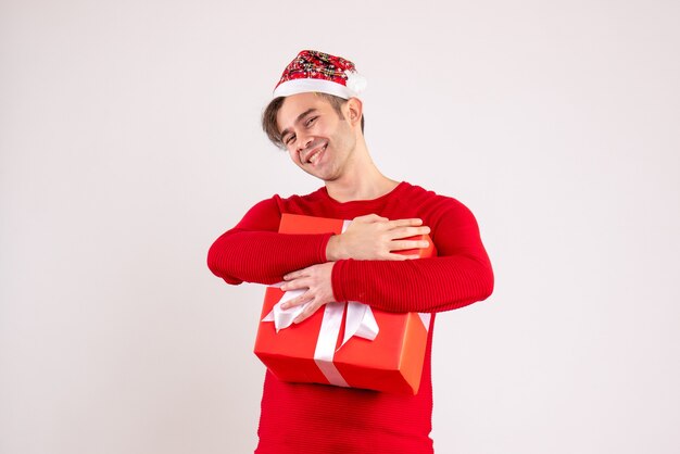 Front view smiling young man with santa hat holding tight his gift on white 