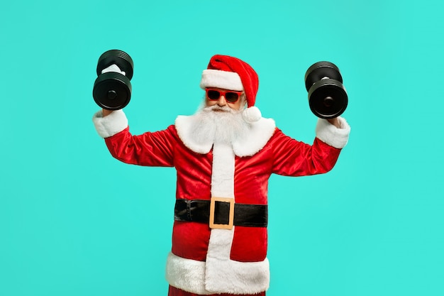 Free photo front view of sportive santa claus holding dumbbels. isolated portrait of funny senior man in christmas costume and sunglasses posing