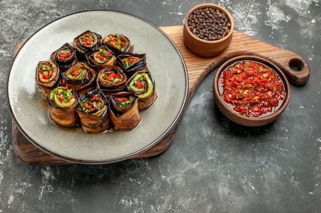 Free Photo front view stuffed aubergine rolls in white oval plate black pepper in bowl on wooden serving board with handle adjika on grey