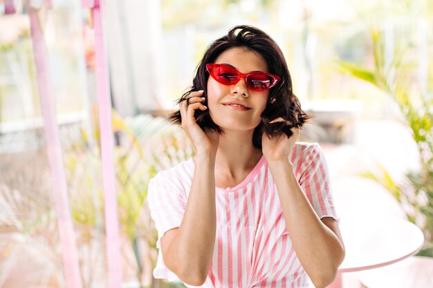 Front view of stunning european girl in striped tshirt Smiling woman in pink sunglasses touching hair on blur background