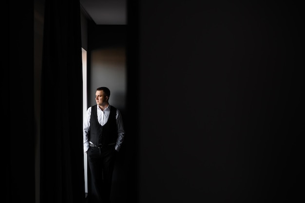 Free photo front view of stylish bridegroom in wedding outfits standing near window keeping hands in pocket and looking away during wedding morning in cozy and dark room