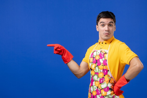 Front view of surprised male housekeeper in apron putting hand on a waist standing on blue wall