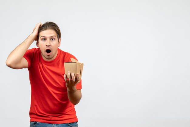 Front view of surprised shocked young guy in red blouse holding small box on white background