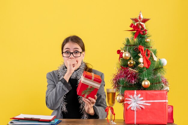 Front view surprised xmas girl with eyeglasses sitting at the table holding xmas gift xmas tree and gifts cocktail