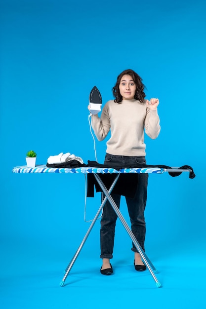 Front view of surprised young woman standing behind the board and holding iron on blue wave surface