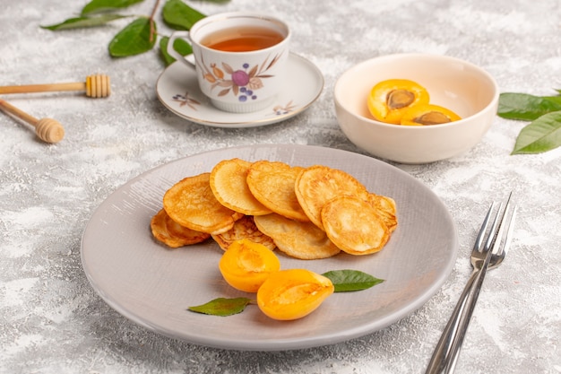 Front view sweet pancakes inside plate with apricots and tea on the grey desk pancake food meal sweet dessert fruit