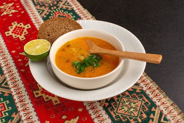 Front view tasty vegetable soup inside plate with bread loaf and lemon on dark desk.