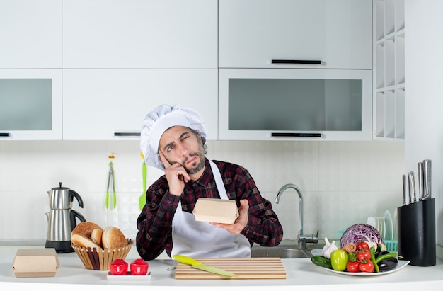 Front view thinking male chef holding burger box in the kitchen