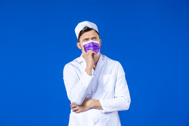 Front view of thinking male doctor in medical suit and purple mask on blue 