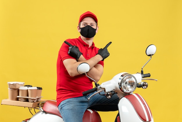 Front view of thinking young adult wearing red blouse and hat gloves in medical mask delivering order sitting on scooter pointing both sides on yellow background