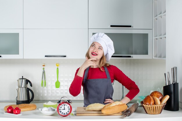 Front view thinking young woman in cook hat and apron putting hand on her waist in the kitchen