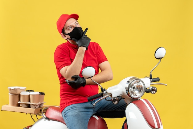 Front view of thoughtful young adult wearing red blouse and hat gloves in medical mask delivering order sitting on scooter on yellow background