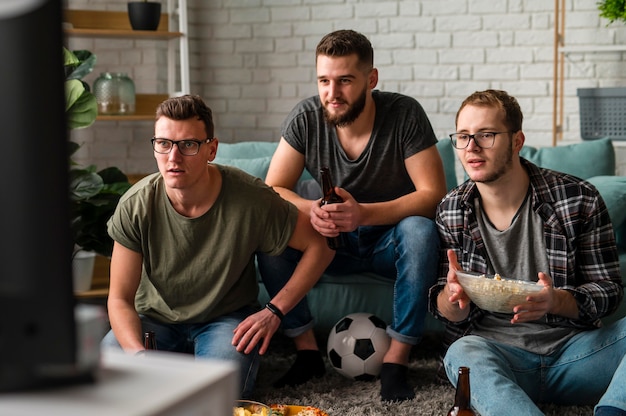 Free photo front view of three male friends watching sports on tv together while having beer and snacks