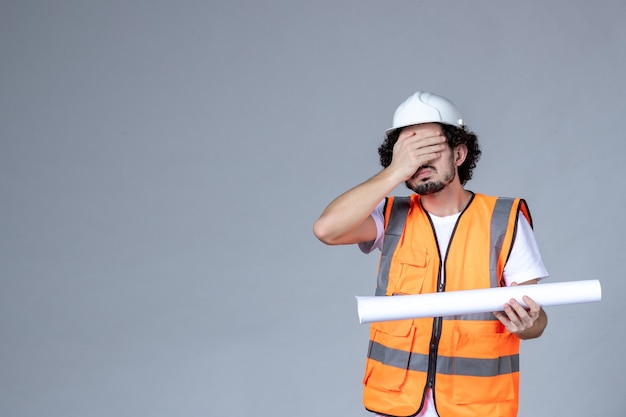 Front view of tired male constructor in warning vest wearing safety helmet and holding blank on gray wall