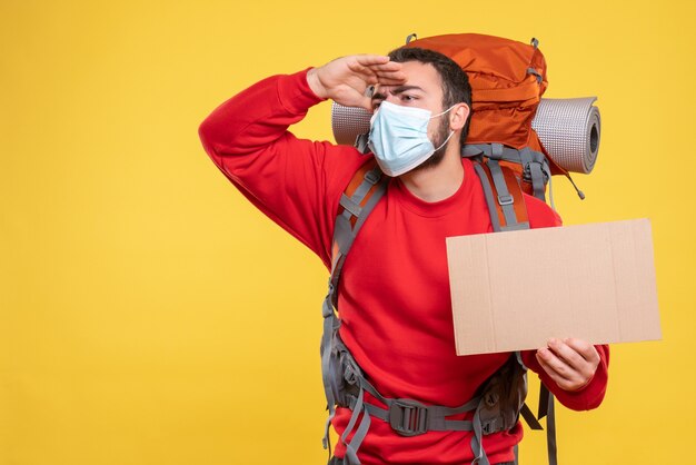 Front view of traveller guy wearing medical mask with backpack showing a sheet without writing looking at something carefully on yellow background