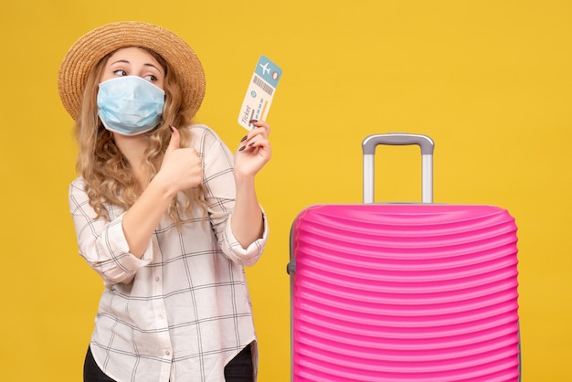 Free photo front view of travelling girl wearing mask showing ticket and standing near her pink bag making ok gesture on yellow