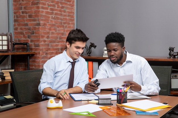 Front view two businessmen sitting at desk one of them using stapler