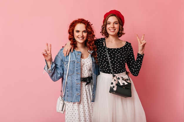 Front view of two friends embracing on pink background. Studio shot of smiling girls showing peace signs.