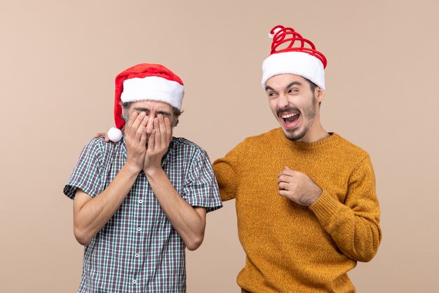 Front view two guys with santa hats one covering his face with hands and the other laughing on beige isolated background