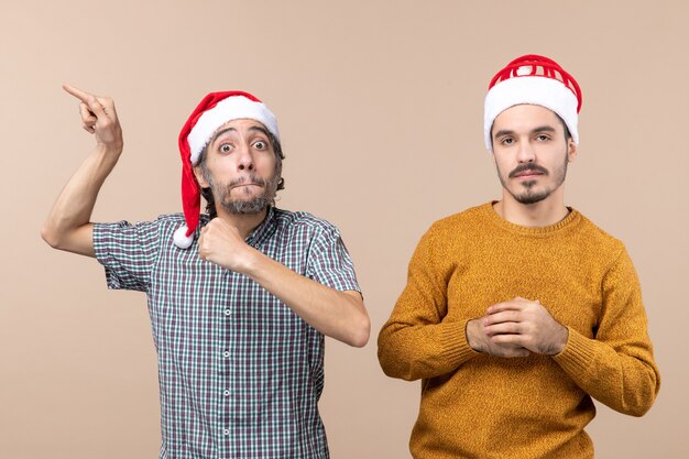 Front view two guys with santa hats one showing something with great interest while standing on beige isolated background