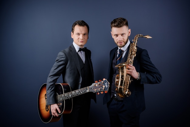 Front view of two men holding musical instruments guitar and saxophone looking at the camera