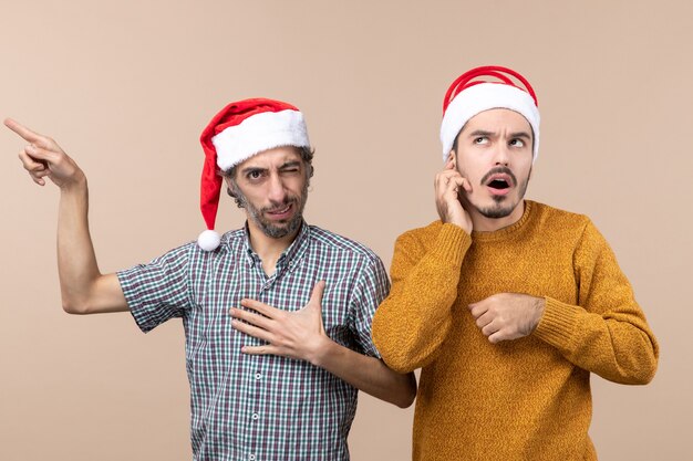 Front view two xmas guys with santa hats one showing something the other thinking on beige isolated background