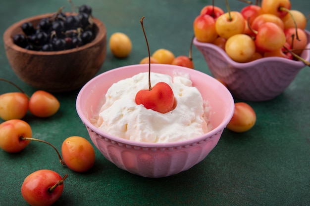 Front view of whipped cream with black currants and white cherries in bowls on a green surface