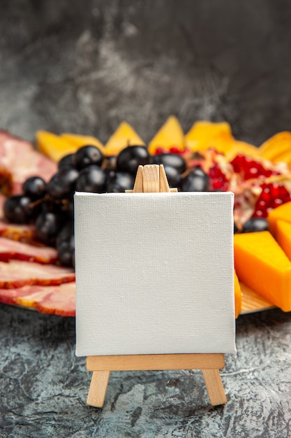 Free photo front view white canvas on wooden easel grapes cheese pieces meat slices on wood plate on dark