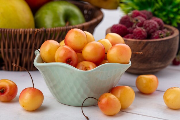 Front view of white cherries in a bowl with raspberries on a white surface