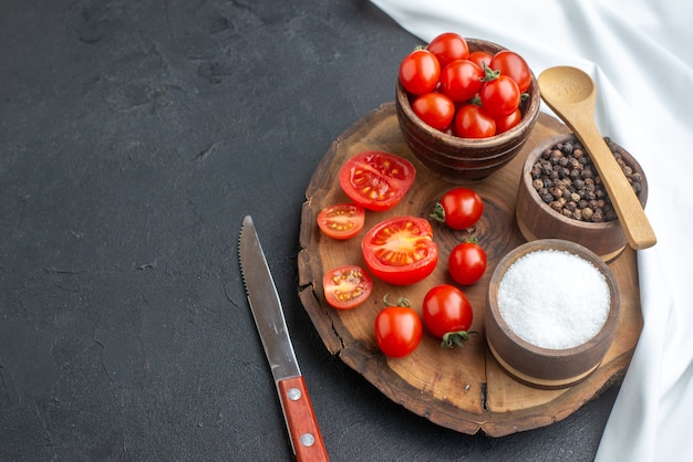 Front view of whole cut fresh tomatoes and spices on wooden board white towel knife on the left side on black surface with free space