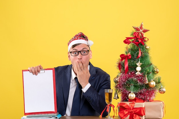 Front view of wide-eyed business man holding clipoard sitting at the table near xmas tree and presents on yellow